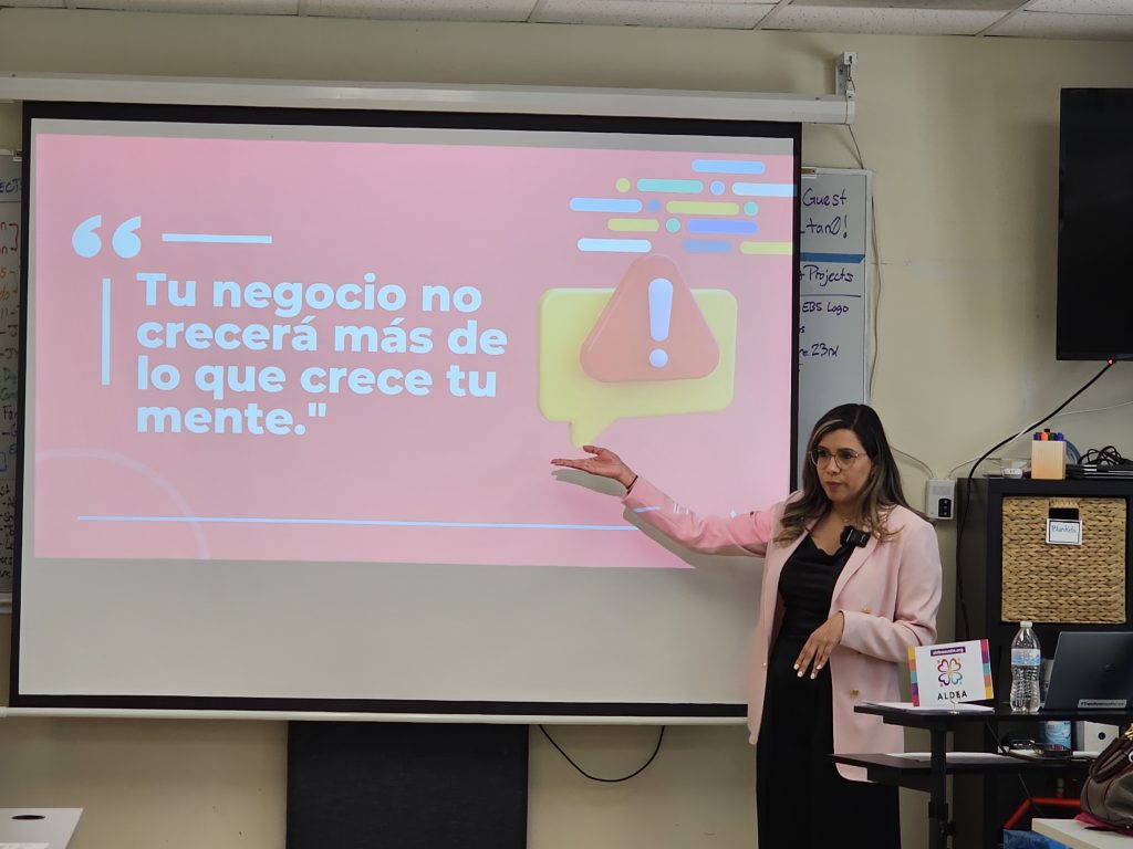 Una mujer latina realiza una presentación en PowerPoint sobre cómo ser propietaria de un negocio. En la pantalla se lee: “Tu negocio solo crecerá tanto como crezca tu mente”.