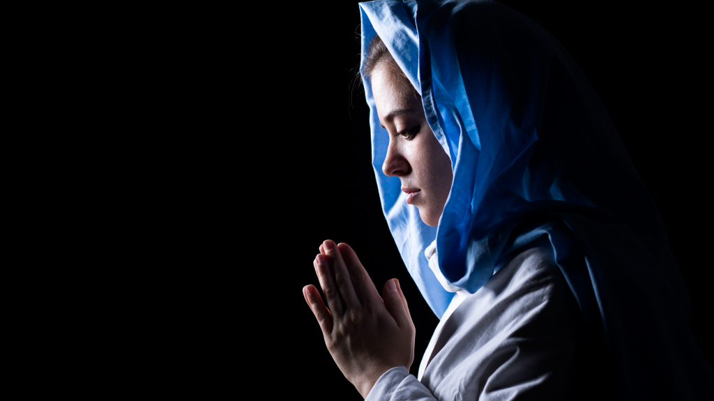 A woman resembling the Virgin Mary wearing a blue satin head covering and holding her hands in prayer.