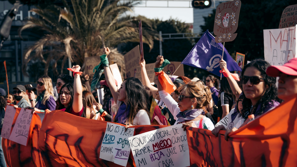 Women at a women's rights March in Latin America hold signs and a giant banner, one sign reads “Quiero dejar de tener miedo cuando mi hija sale de casa”